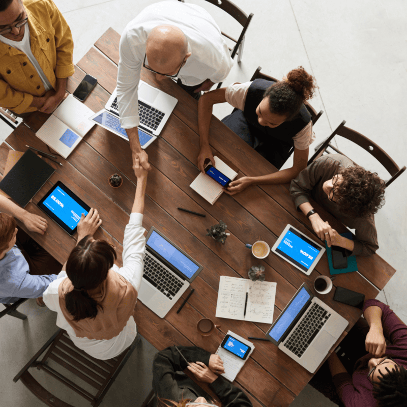 View from above of a group of meeting attendees around a table, with laptops, pads and pencils.